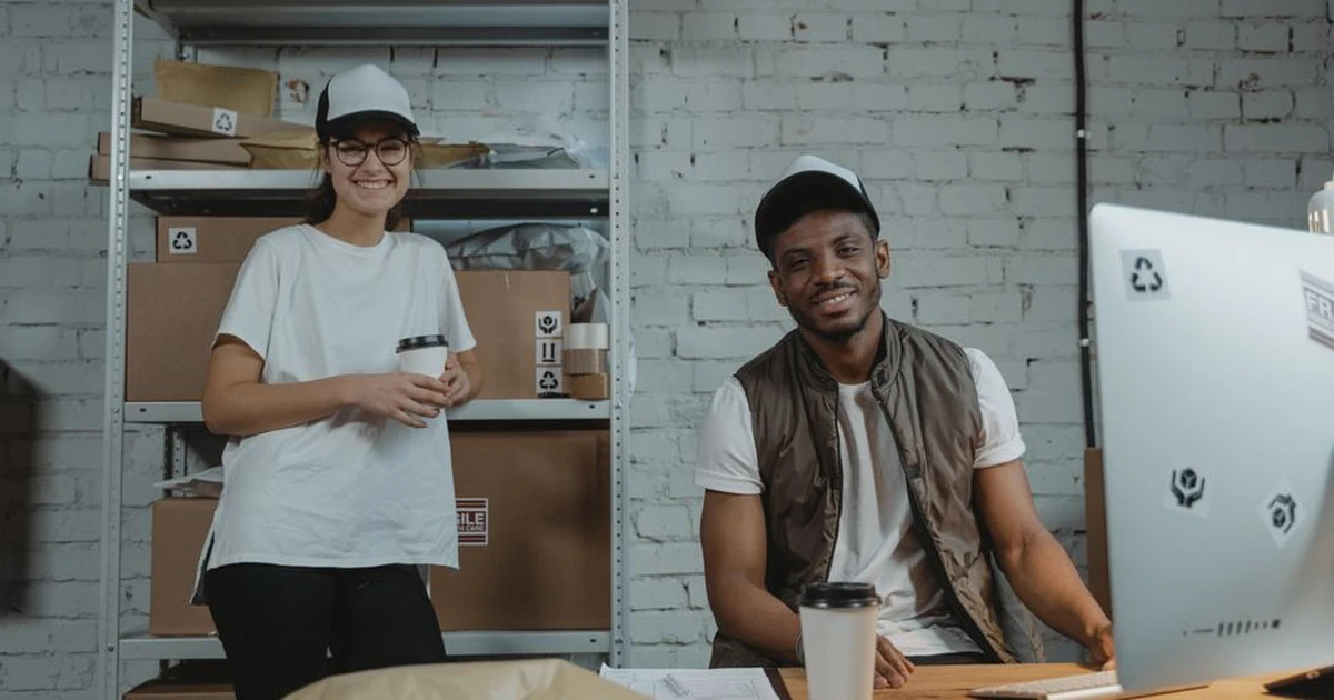 Two warehouse employees in casual attire smiling while working in a courier environment with packages.