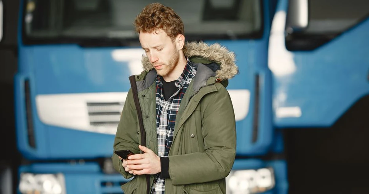 A man wearing a parka checks his mobile phone in front of a blue truck parked in a garage.