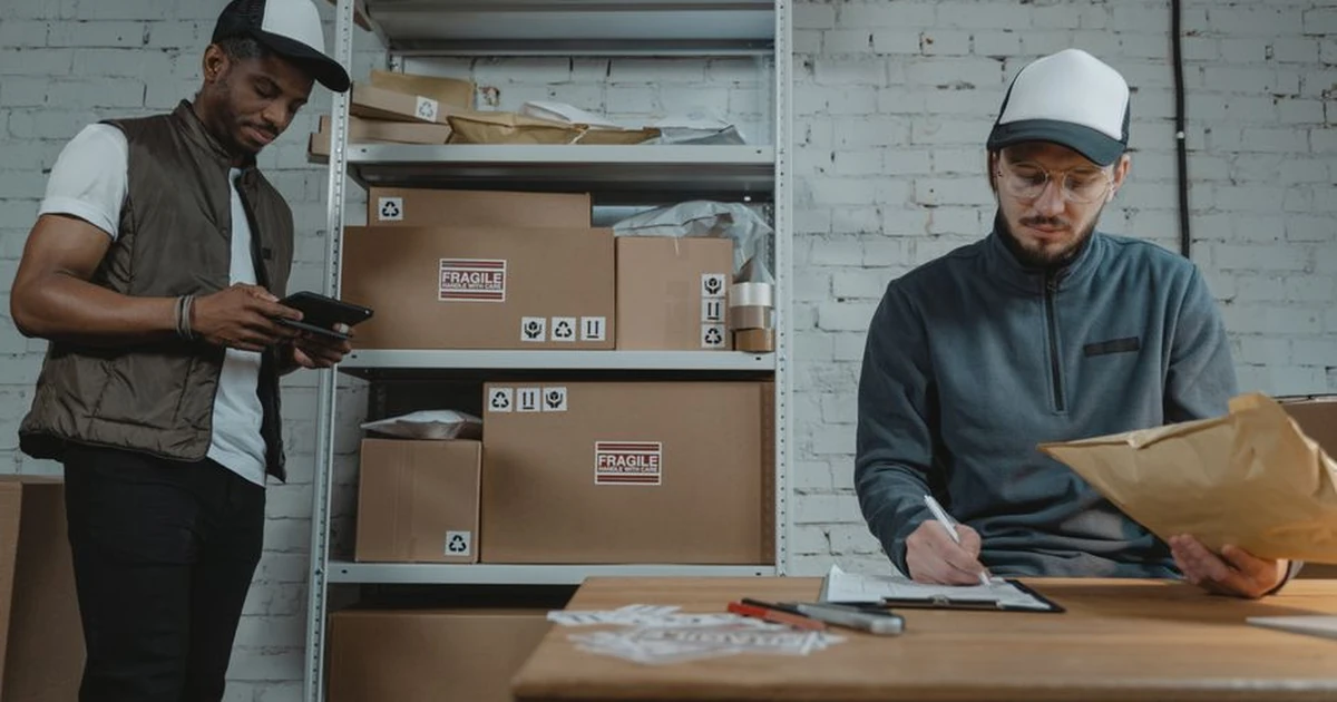 Warehouse employees organizing shipments with packages and documentation in a brick-walled workspace.